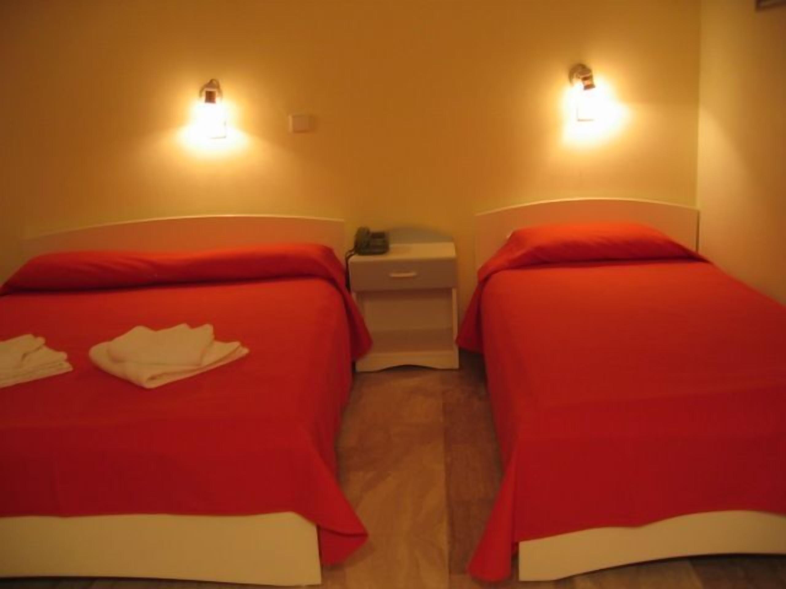 A hotel room with two single beds, each with a red bedspread and white pillows. There are two bedside tables, one on each bed, with a lamp on each. The room has a warm, yellowish lighting.