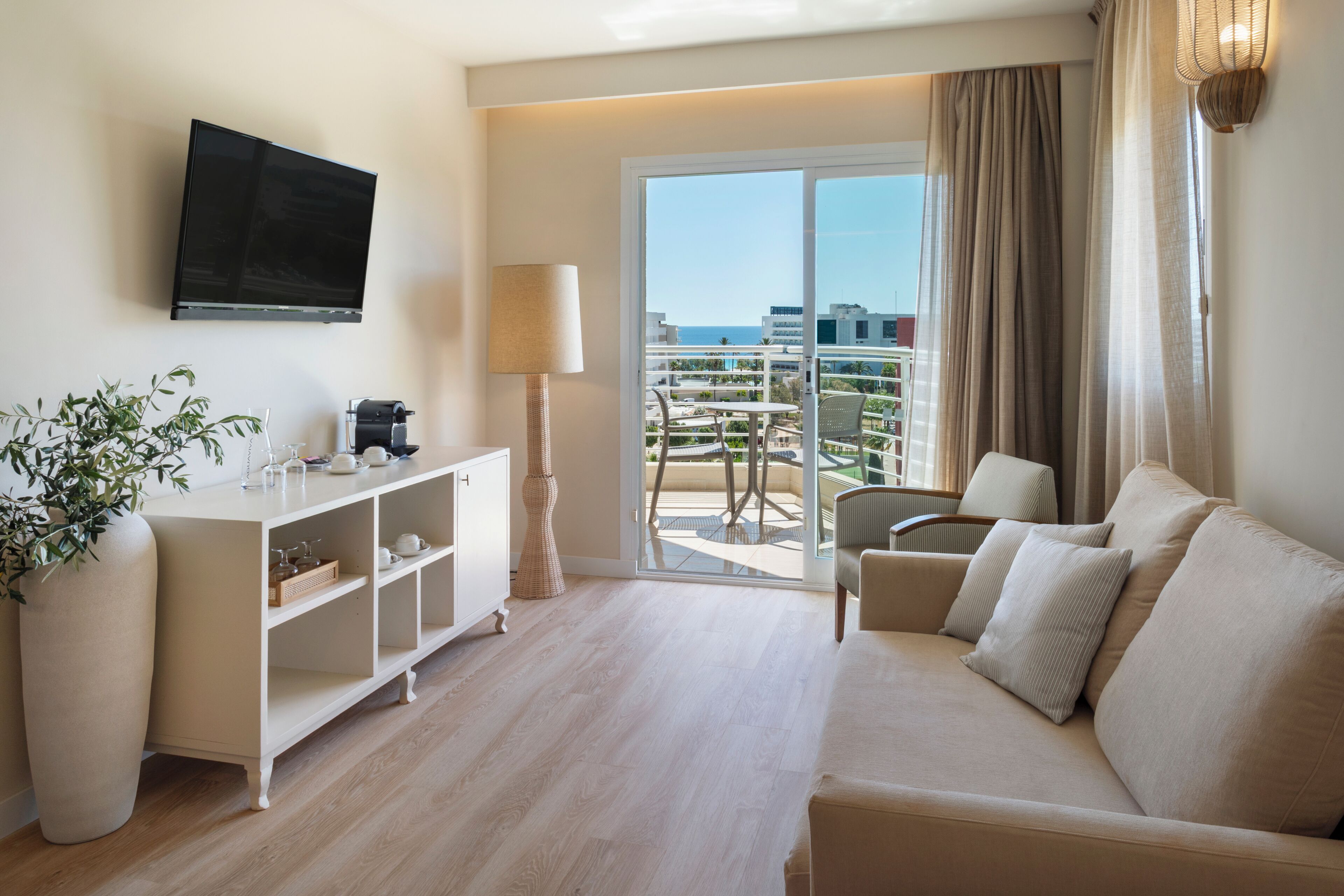 Hotelkamer met een bank, televisie, plant, lamp en balkon met uitzicht op de stad en de zee.
