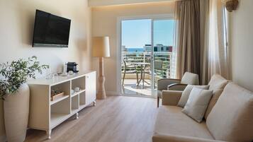 Hotelkamer met een bank, televisie, plant, lamp en balkon met uitzicht op de stad en de zee.