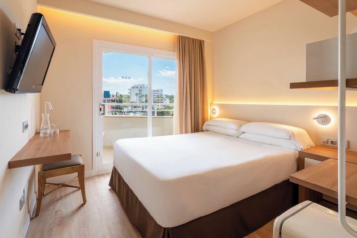 Ein Hotelzimmer mit Bett, Nachttisch, Fernseher und Blick auf Gebäude durch das Fenster.