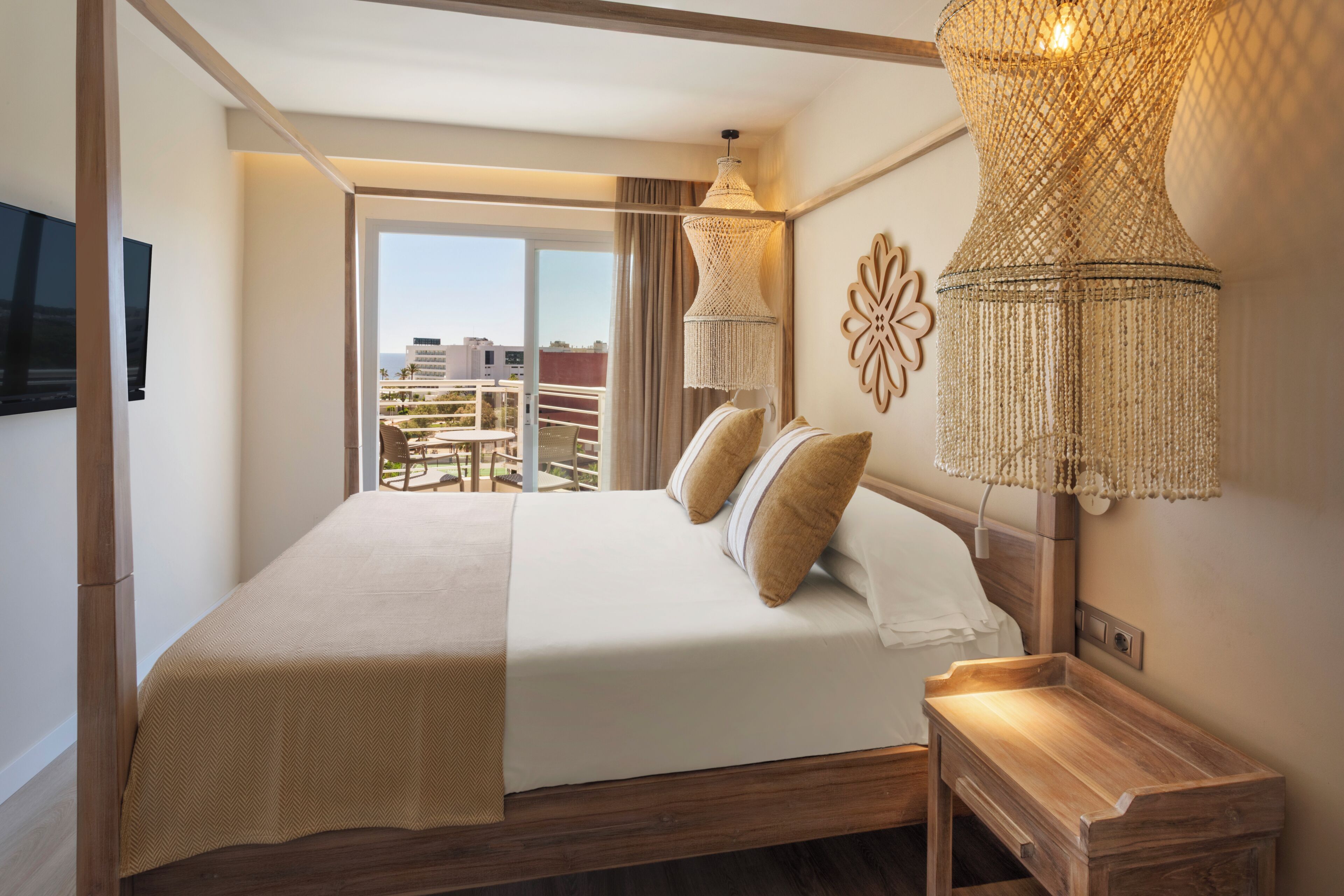 A hotel room with a bed, a wooden bedside table, a large woven lamp, and a view of the outdoors.