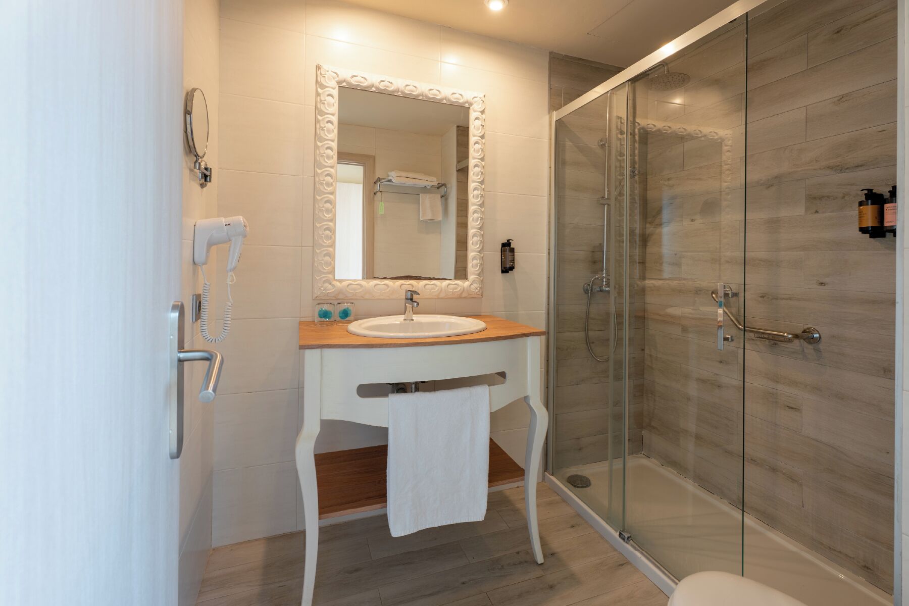 A modern bathroom with a glass-enclosed shower, a white sink with a wooden countertop, and a large mirror with an ornate frame.