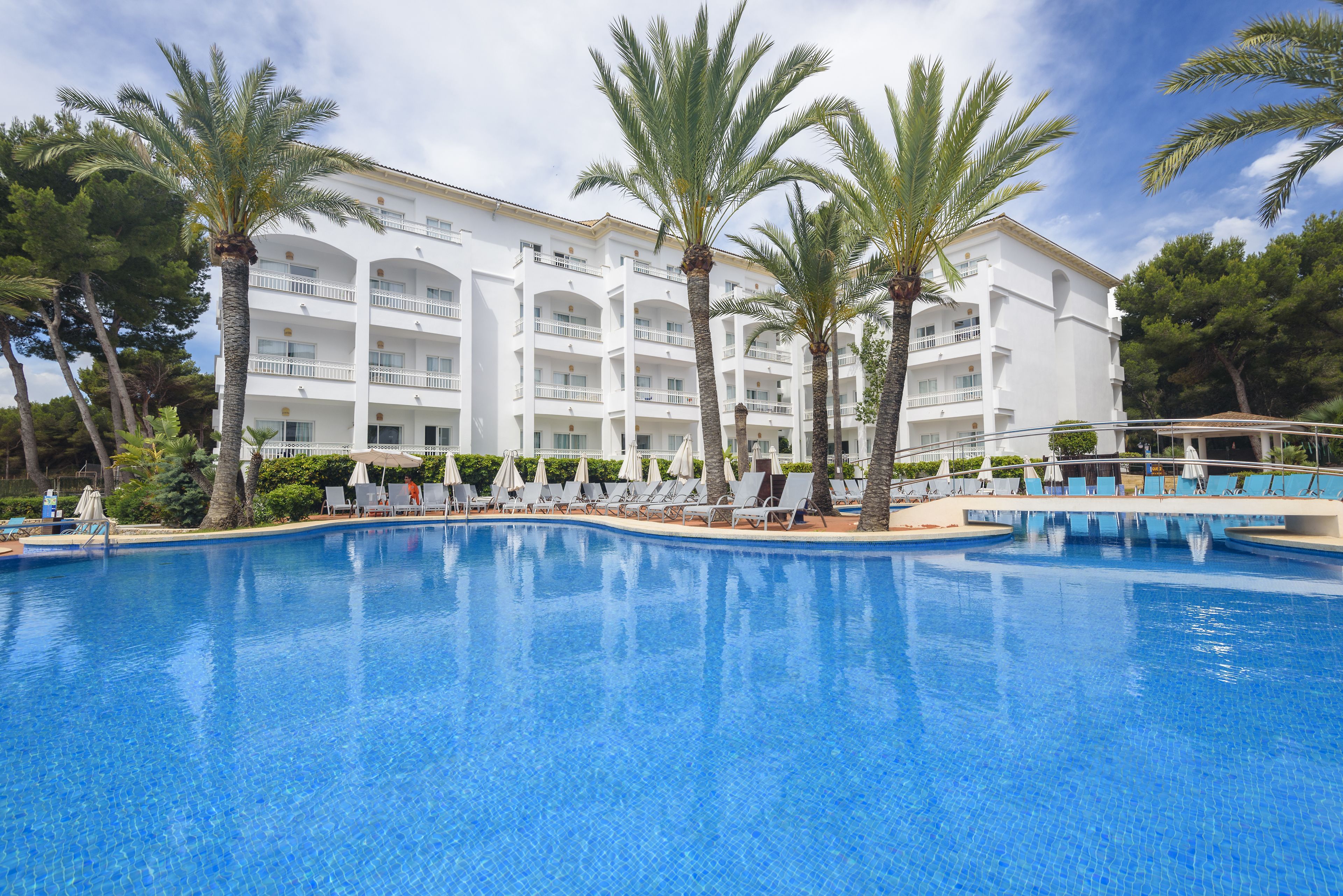 Un hôtel doté d’une piscine, de palmiers et d’un bâtiment blanc orné de nombreux balcons.