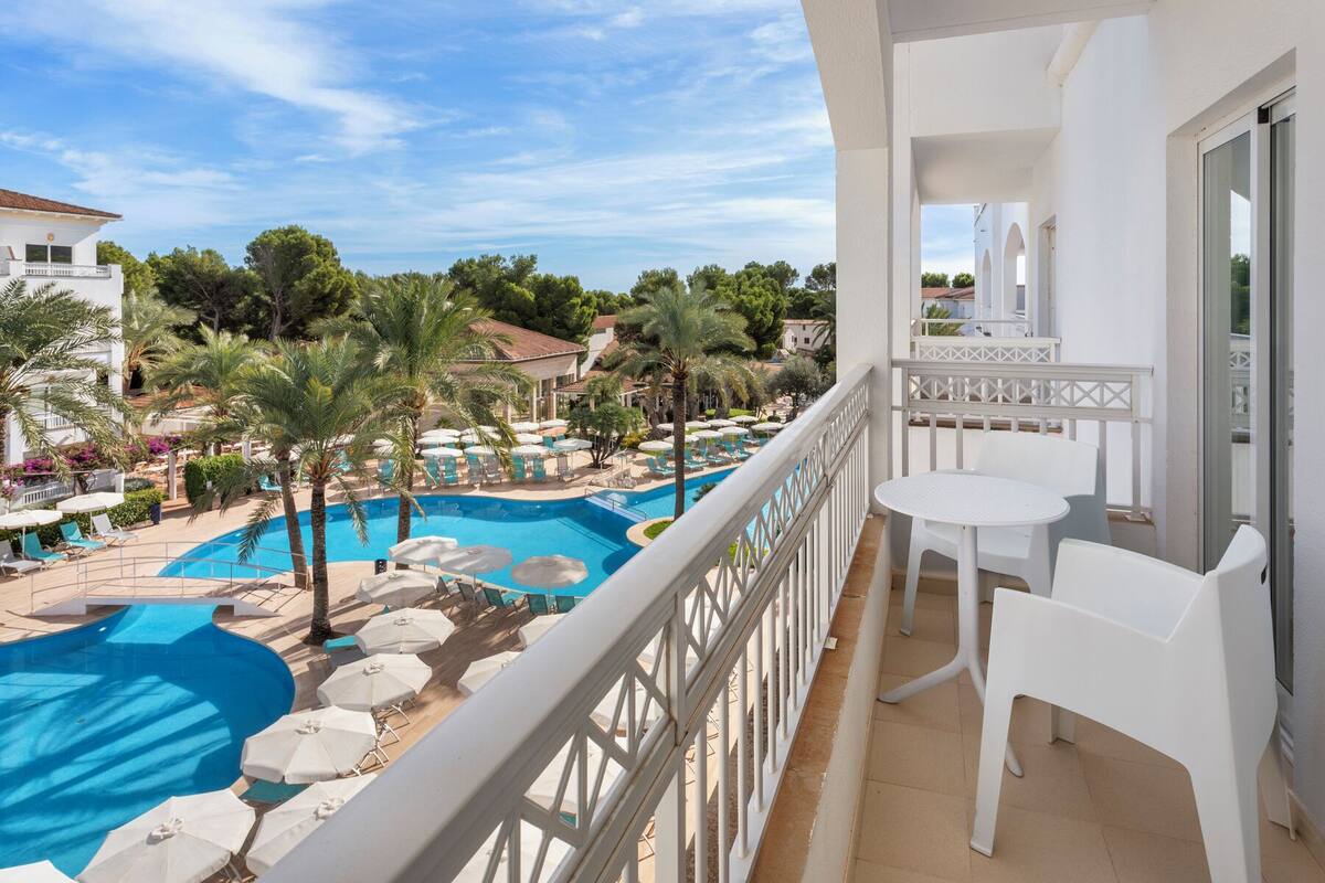 Balcón con vista a la piscina, muebles blancos y cielo despejado.