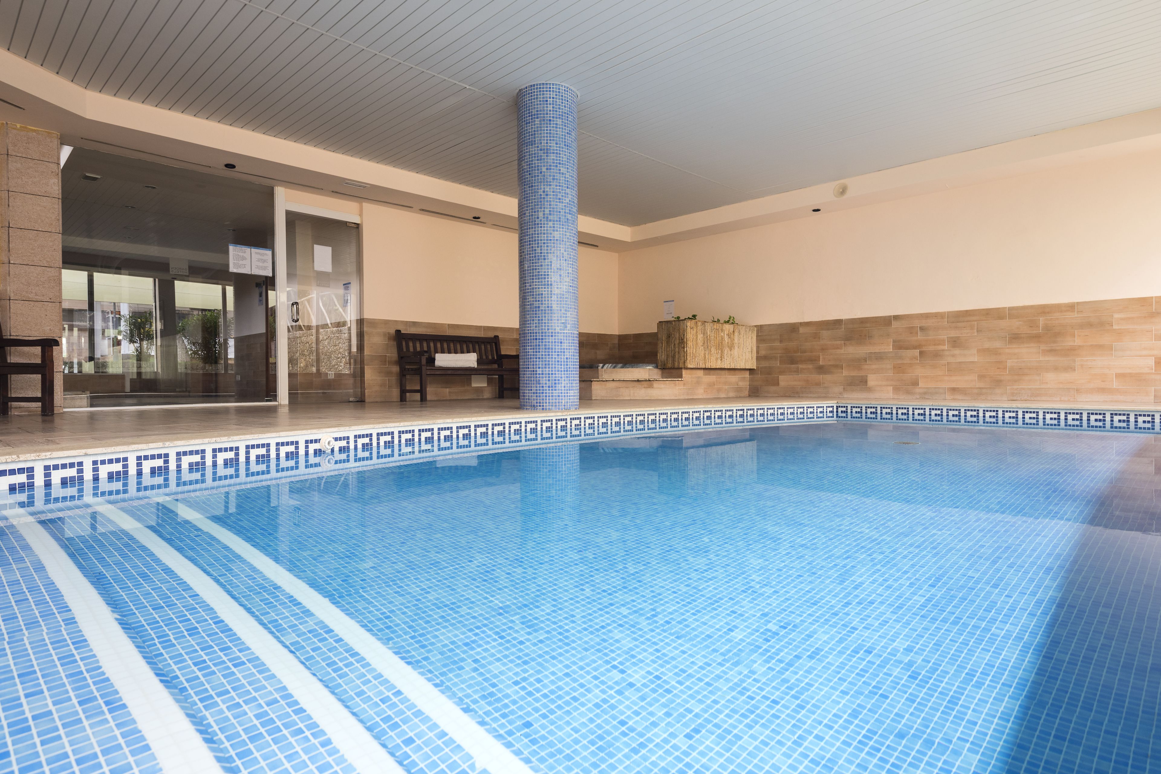 Una piscina con baldosas azules y blancas, un banco de madera y un pilar revestido de baldosas.