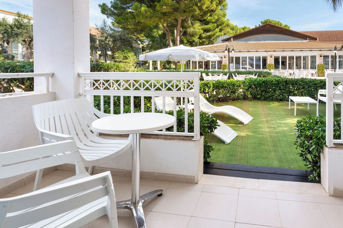 Un patio con mobiliario blanco, una pequeña mesa redonda y una silla blanca. Hay una sombrilla blanca y una silla con respaldo curvo. El patio tiene suelo de baldosas y está rodeado de vegetación y un edificio con tejado de cerámica.