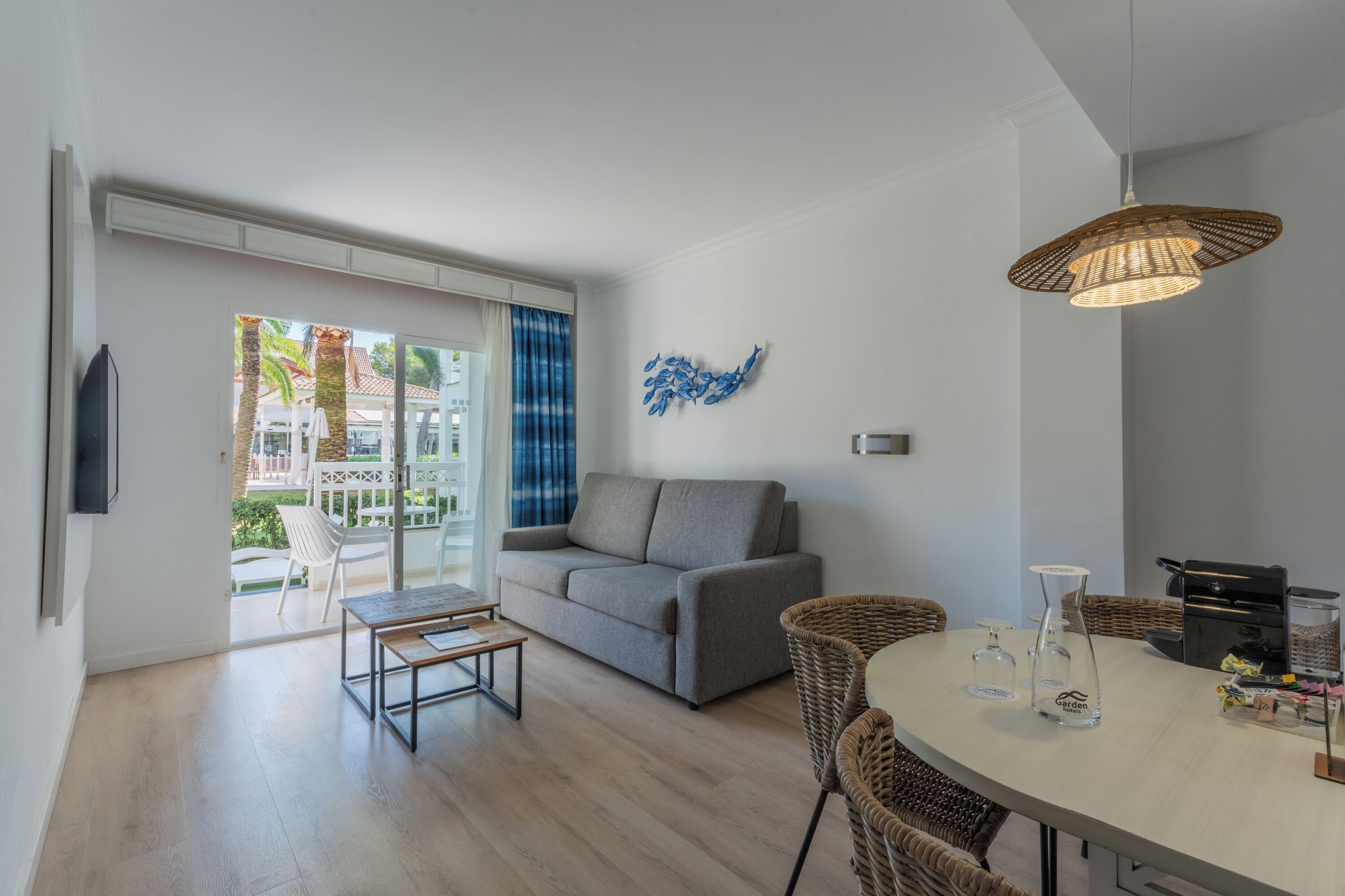 Una sala moderna con un sofá gris, una mesa de comedor con sillas de mimbre y un ventanal con vistas a palmeras.