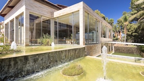A modern building with large glass windows, a stone facade, and a water feature in front. - Aparthotel Green Garden (Capdepera)