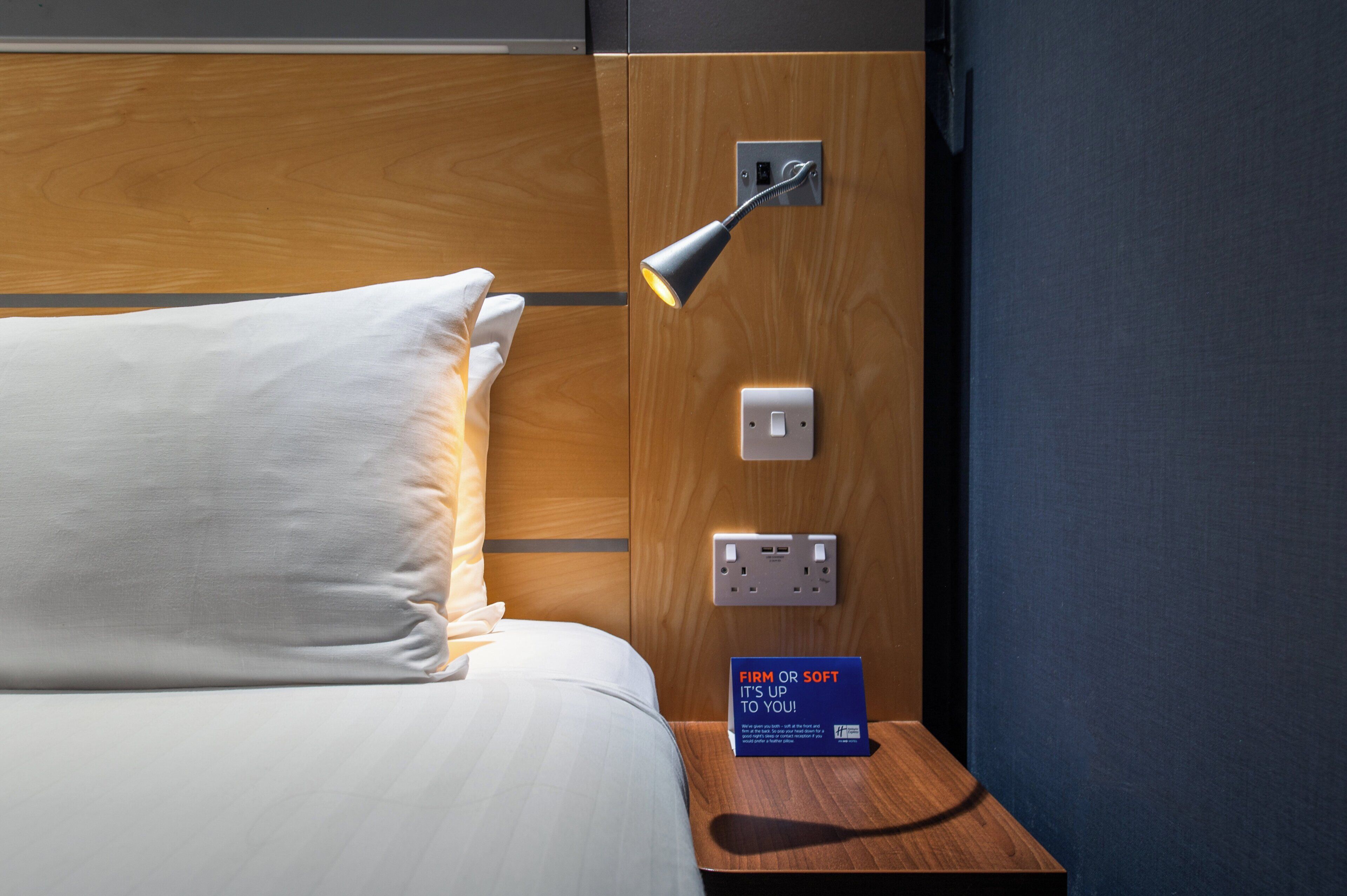 A hotel room with a wooden headboard, a bedside lamp, a power outlet, and a sign indicating the firmness of the mattress.