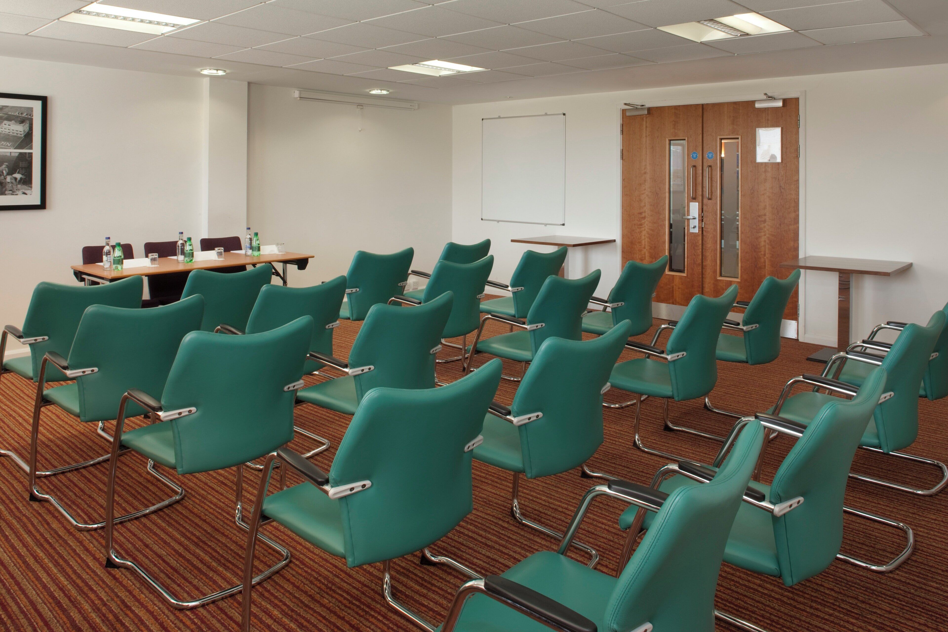 A conference room with rows of teal chairs, a whiteboard, and a wooden door with glass panels.