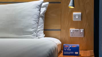 A neatly made bed with a pillow, a bedside lamp, a power outlet, and a sign about pillow firmness.
