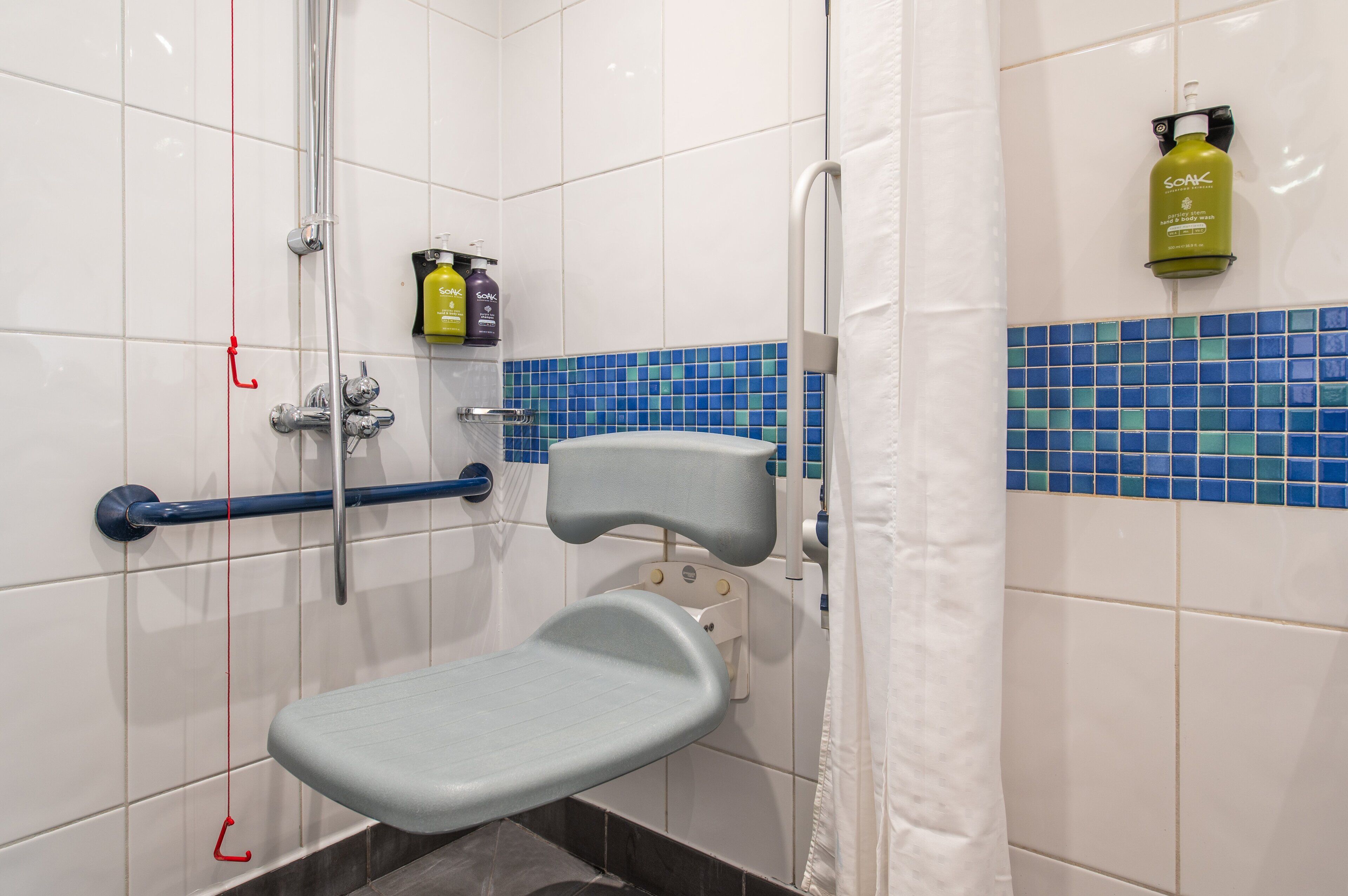 Doccia con maniglione, soffione a mano e sedile per la doccia. Shampoo e balsamo su uno scaffale.