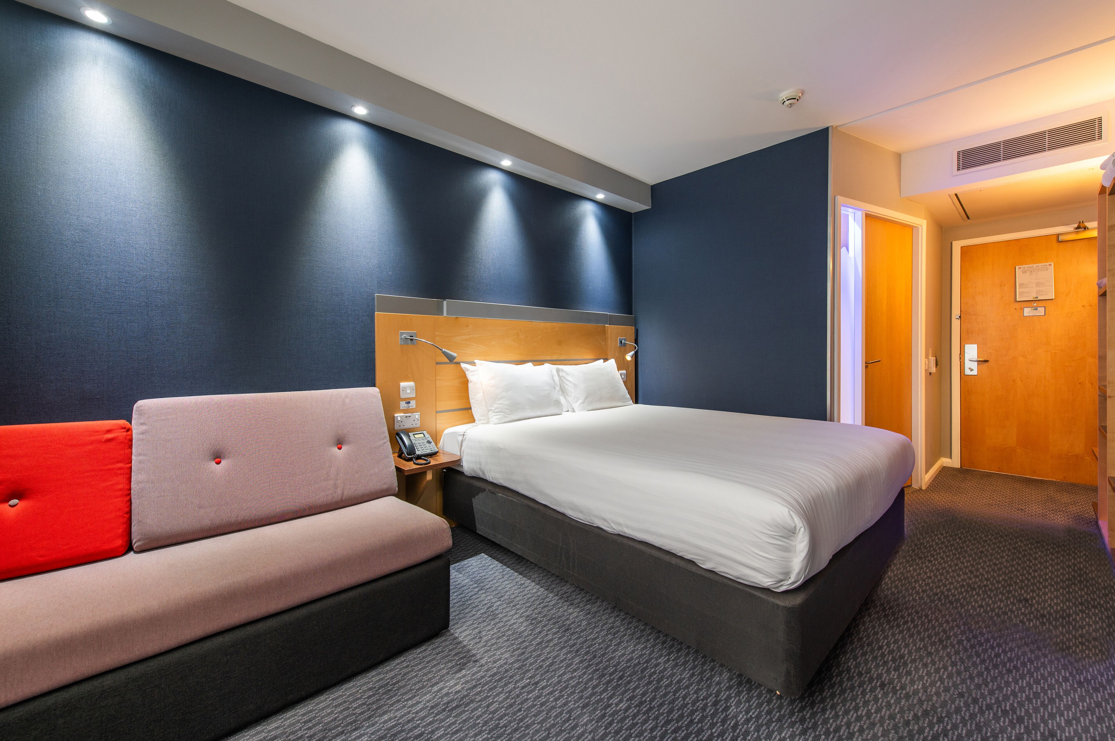 A hotel room with a bed, a sofa, a nightstand, and a door.