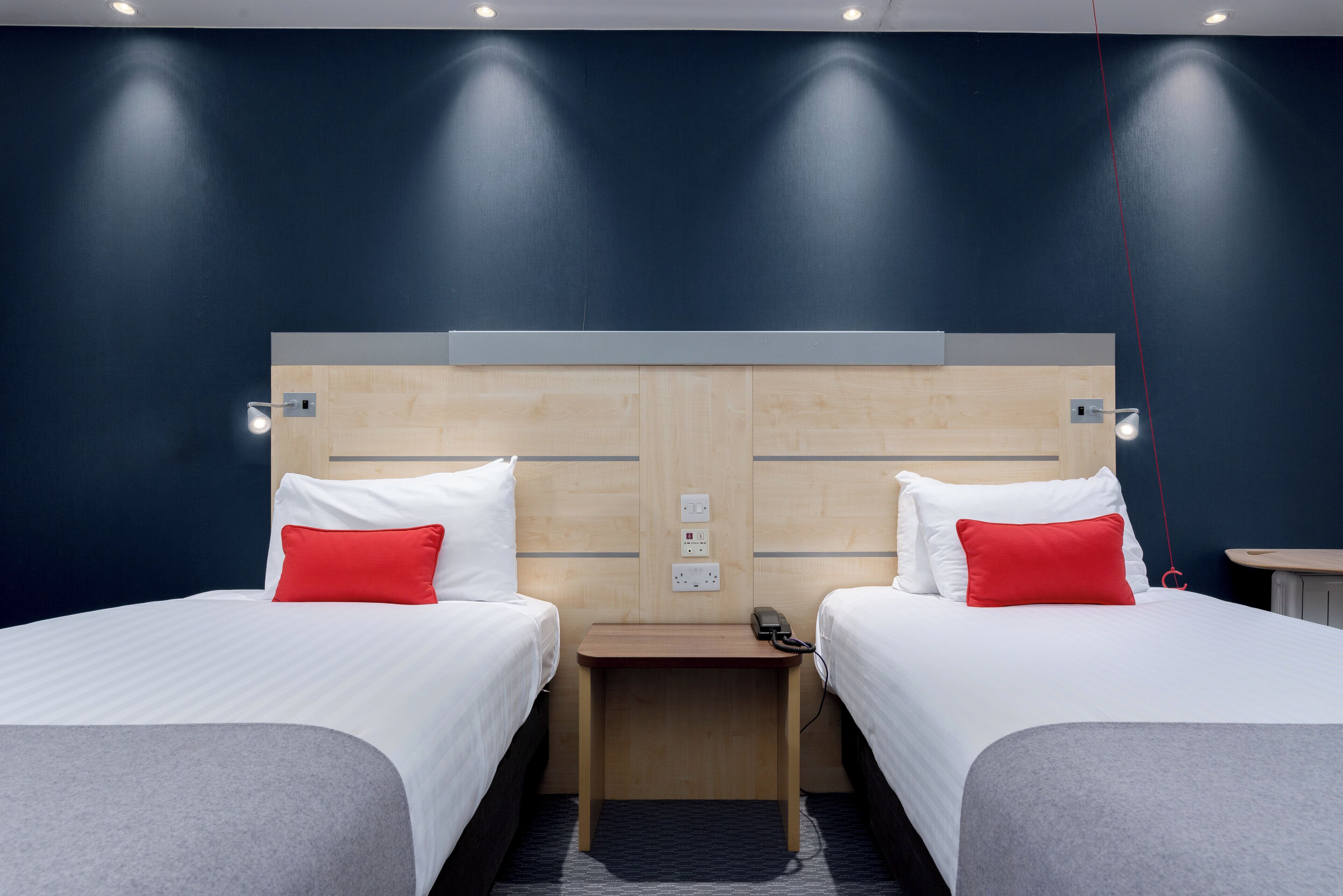 Habitación de hotel con dos camas, cada una con ropa blanca y almohadas rojas, cabecero de madera y una mesita de noche pequeña con un teléfono.
