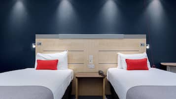 Habitación de hotel con dos camas, cada una con ropa blanca y almohadas rojas, cabecero de madera y una mesita de noche pequeña con un teléfono.