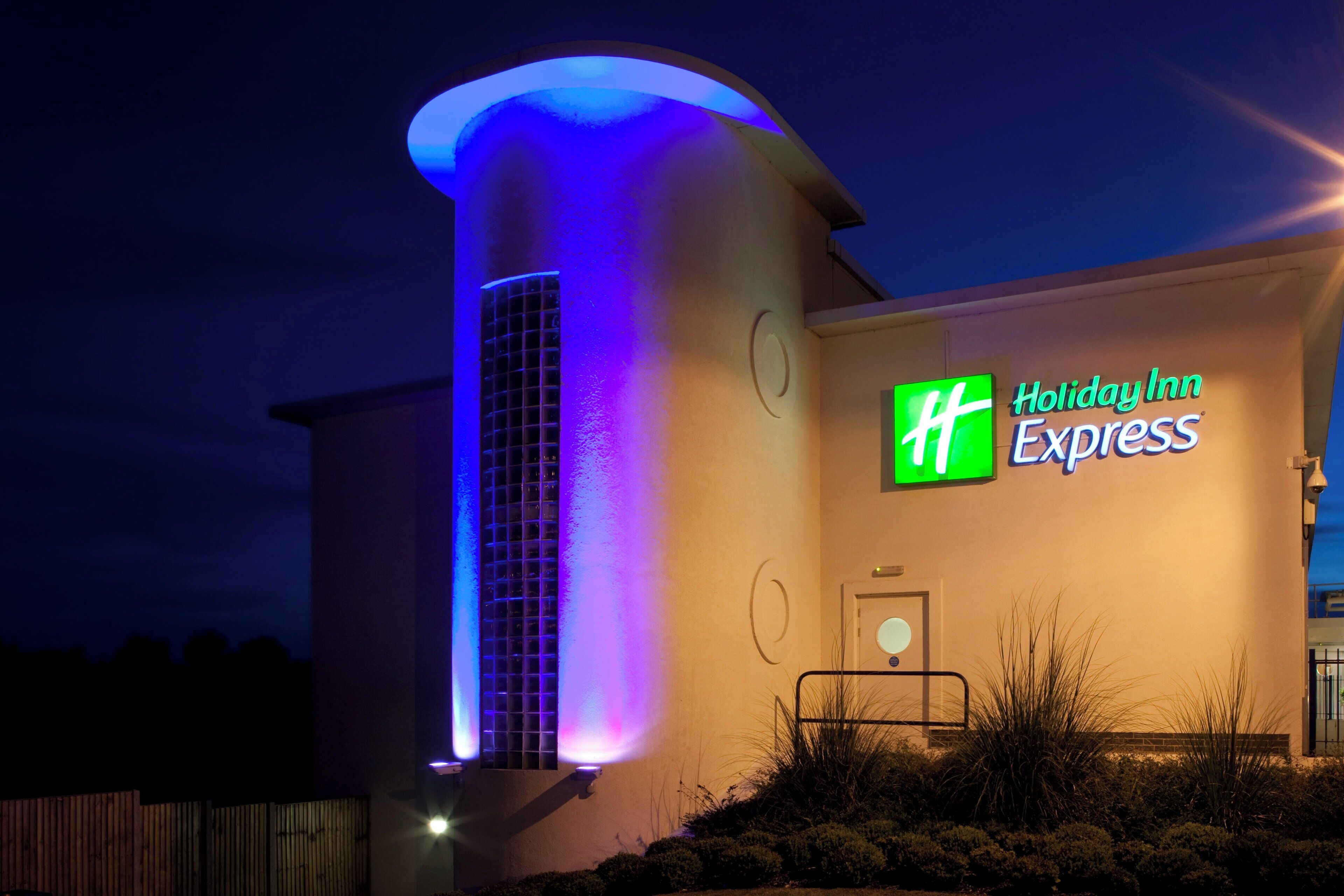 A Holiday Inn Express hotel with a prominent 'HH' sign, a cylindrical structure with a blue-lit entrance, and a flat facade with a door and a small window.