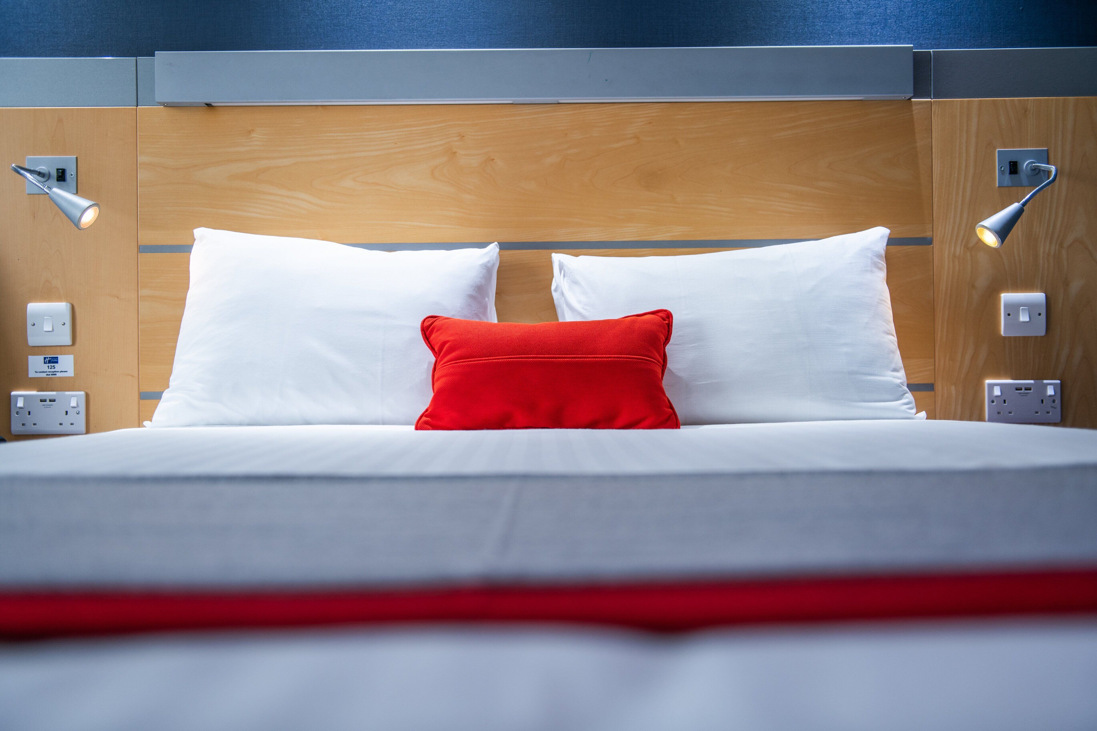 A neatly made bed with white and red pillows, a wooden headboard, and bedside lamps.