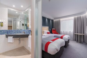 A hotel room with a bathroom, two beds, a mirror, and a window with curtains.