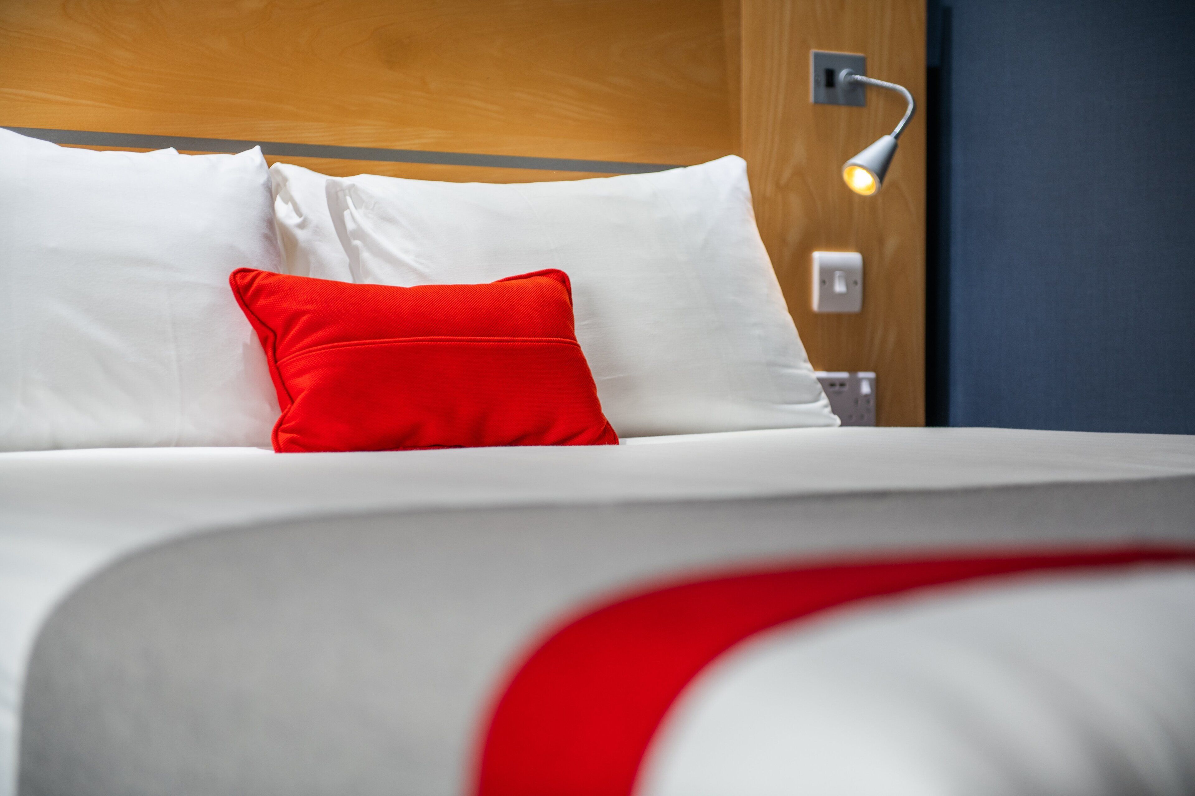 A neatly made bed with white and red pillows, a wooden headboard, and a wall-mounted light fixture.