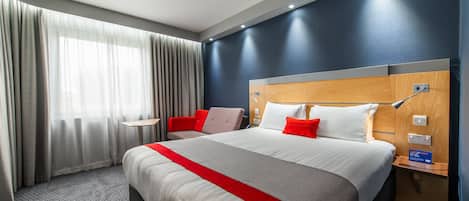 A hotel room with a large bed, a bedside table, a nightstand, a lamp, and a window with curtains.