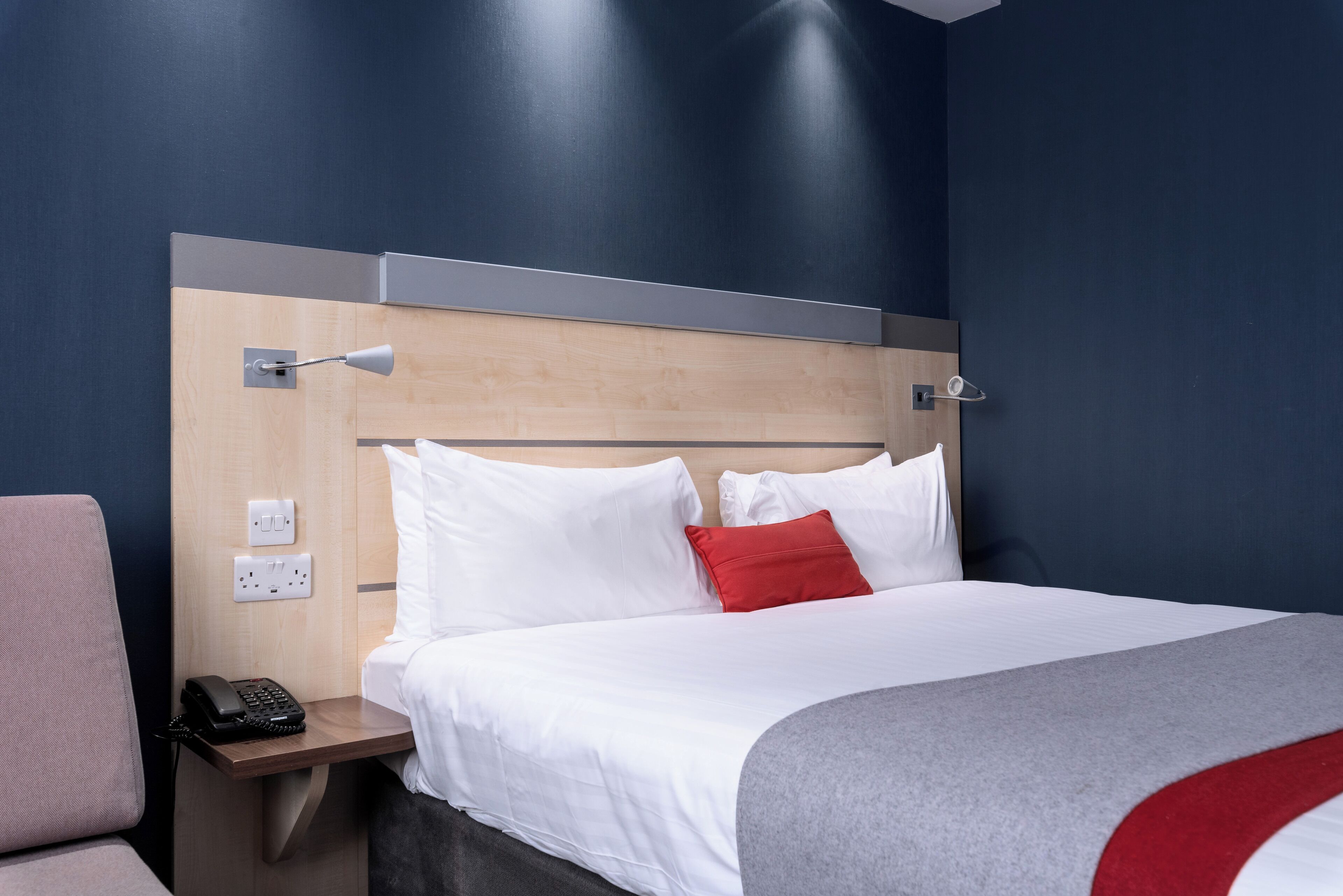 A hotel room with a bed, a nightstand with a phone, and a wall-mounted lamp.