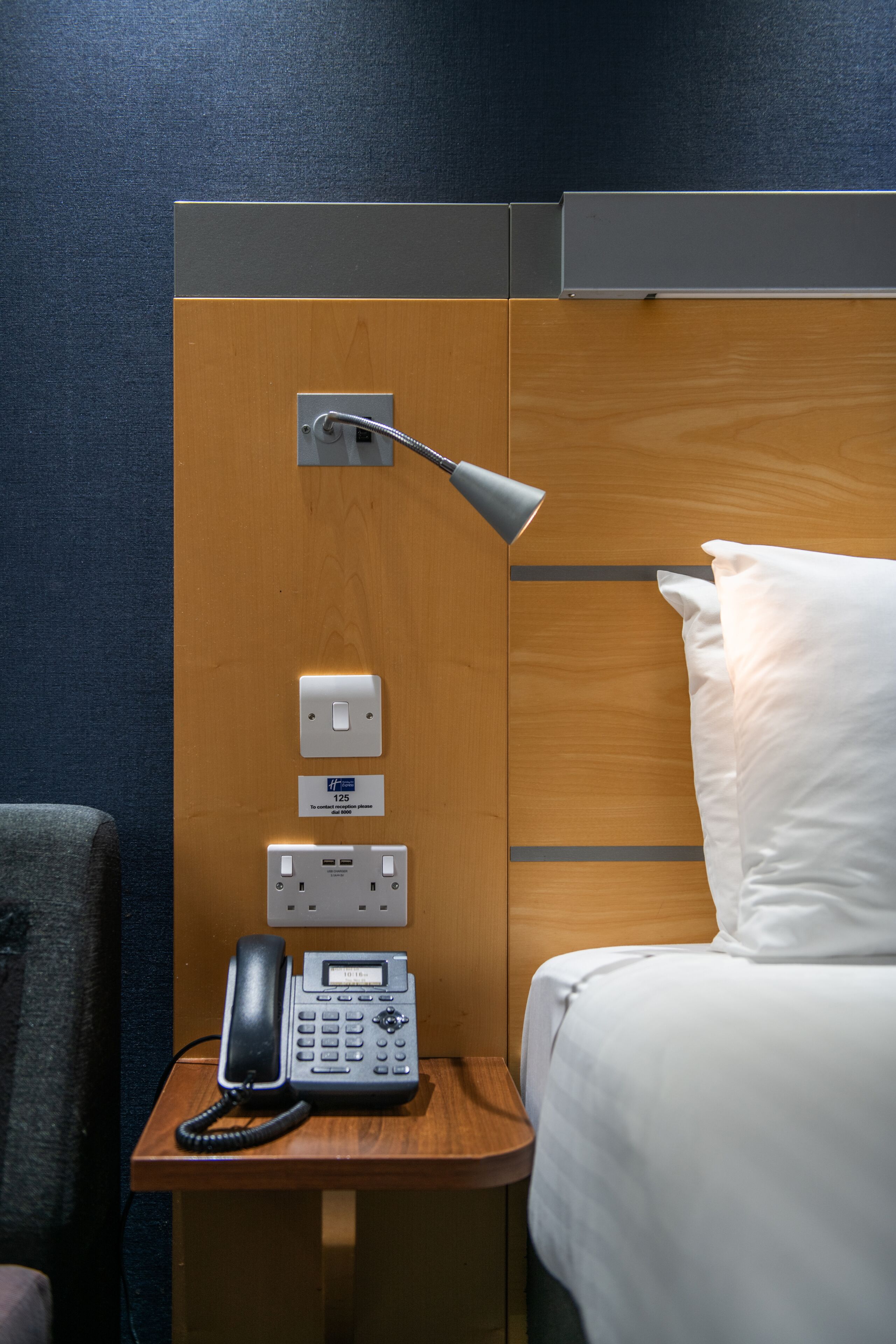 A hotel room with a wooden headboard, a bedside table with a telephone, a lamp, and a power outlet.