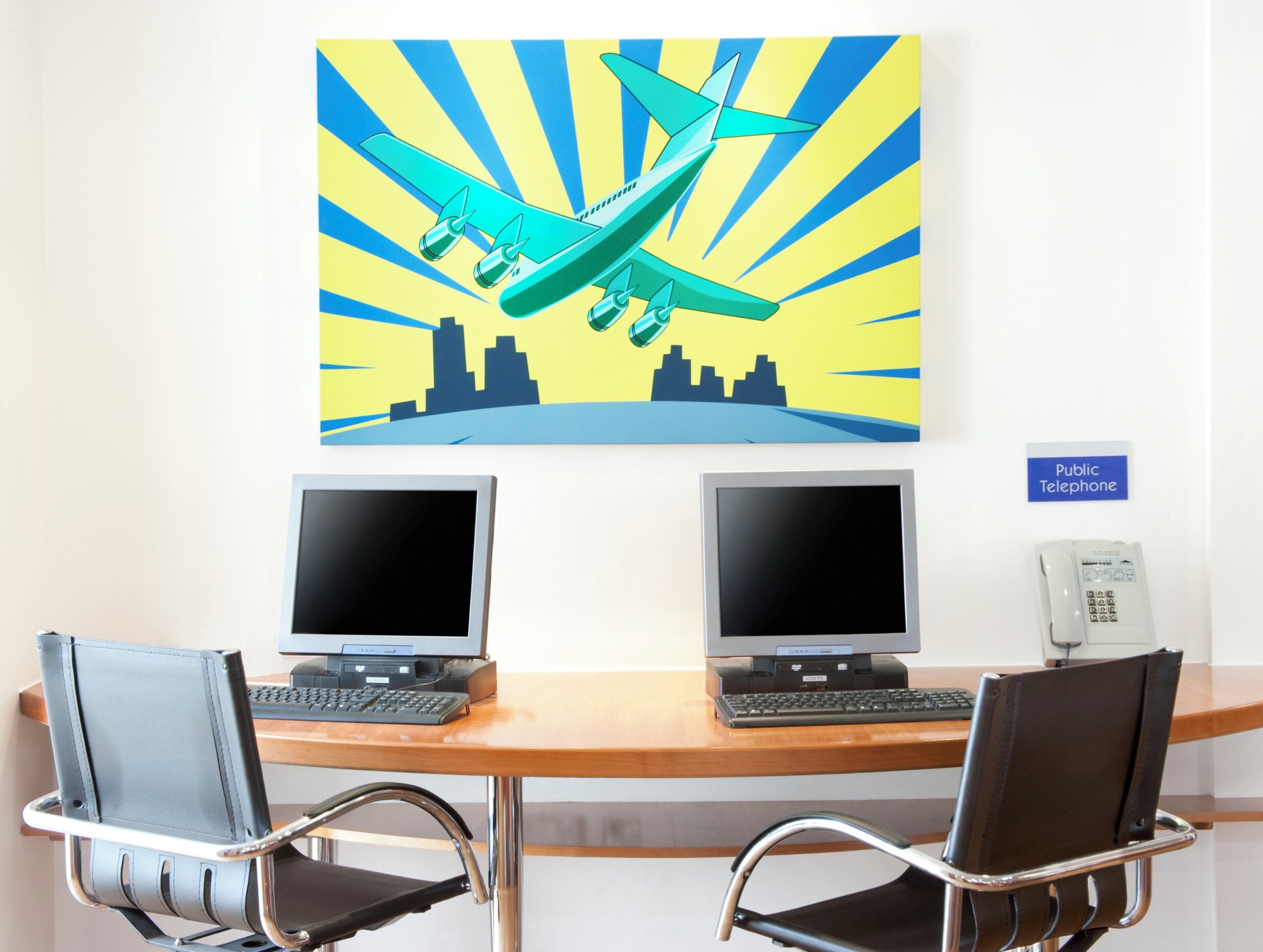 Un espacio de trabajo con dos monitores, un teléfono público y un cuadro de un paisaje urbano.