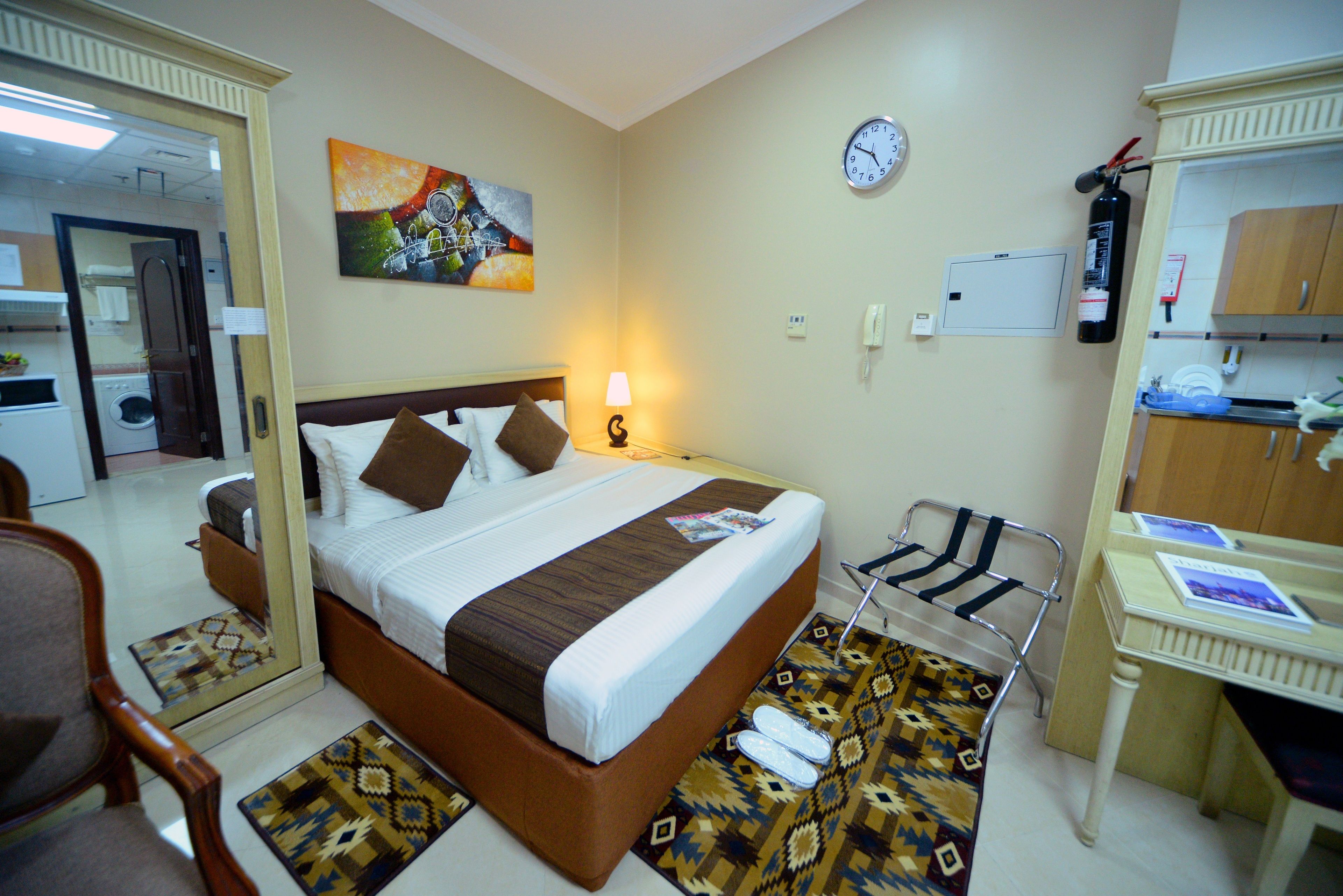 A hotel room with a bed, a desk, a chair, a mirror, a clock, and a fire extinguisher.
