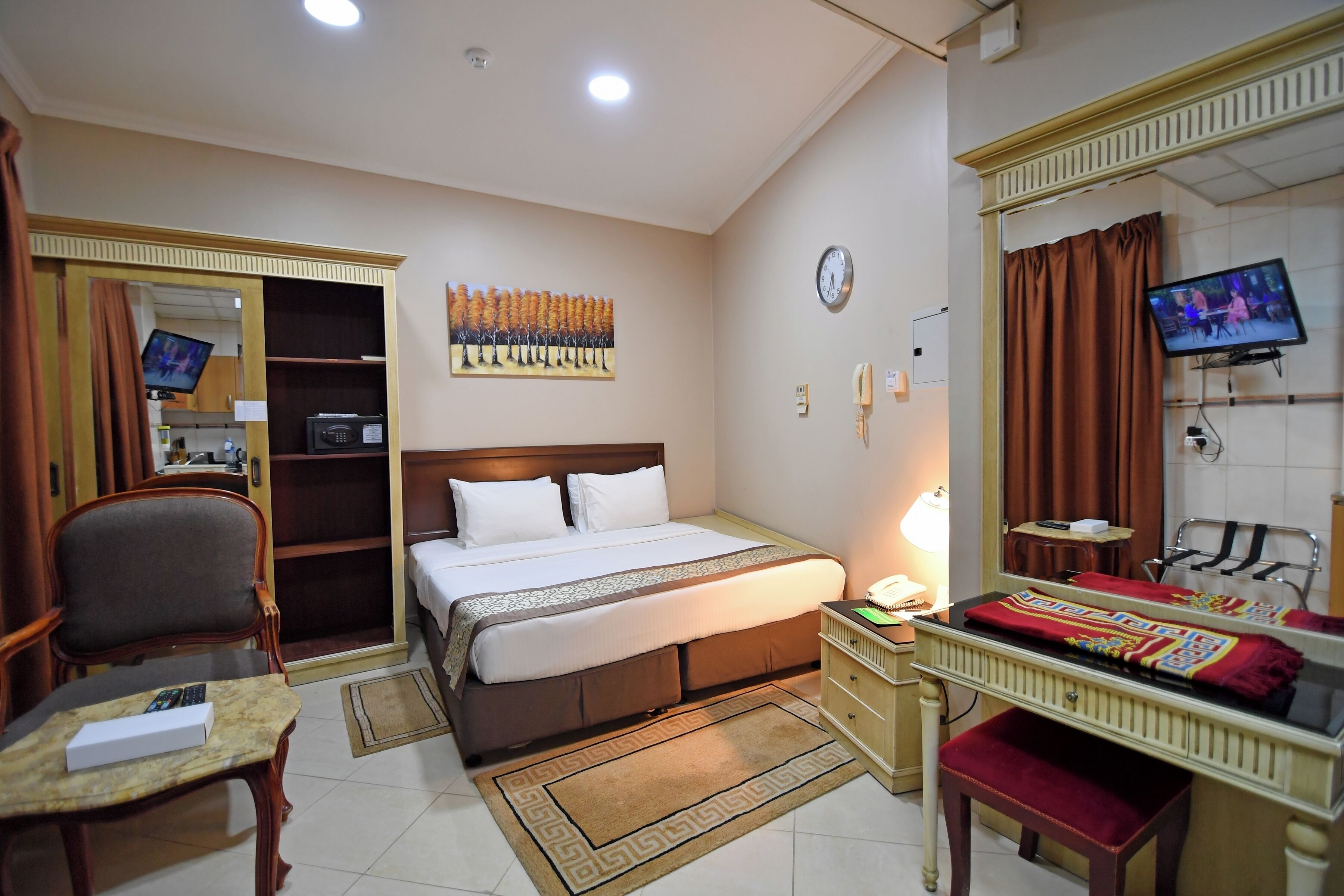 A hotel room with a bed, a desk, a chair, a television, a mirror, and a clock.