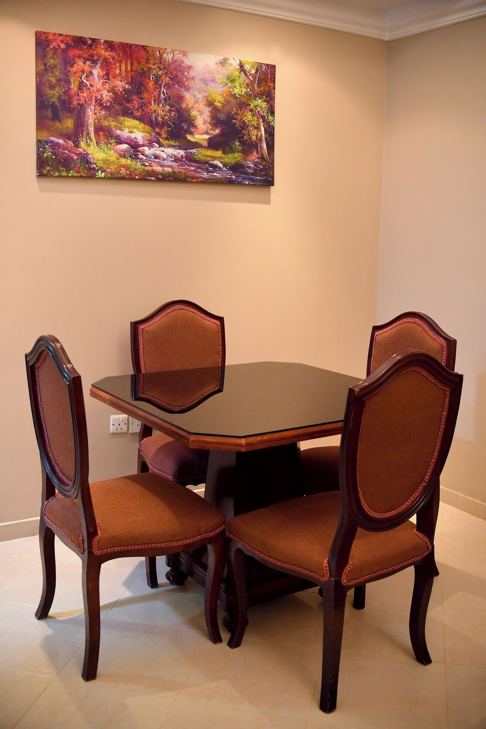 A dining area with a round table and four chairs, a painting on the wall, and a light switch on the wall.