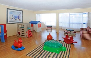 Amplio salón con piso de madera, zona de juegos con juguetes, casita de juegos y un ventanal con cortinas.