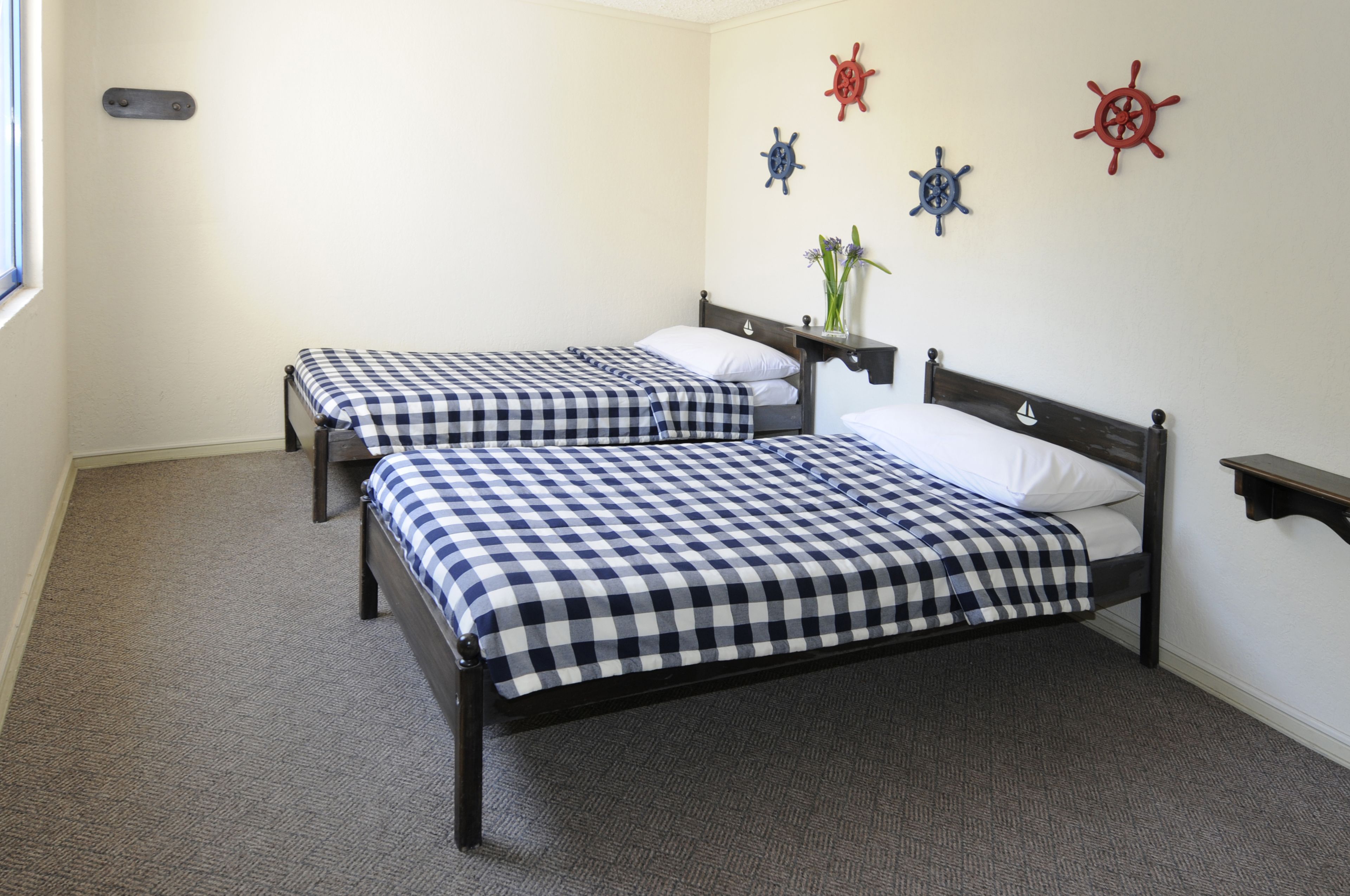 Una habitación con dos camas, cada una con una colcha a cuadros, una mesita de noche con un florero y decoraciones en la pared que parecen timones de barco.