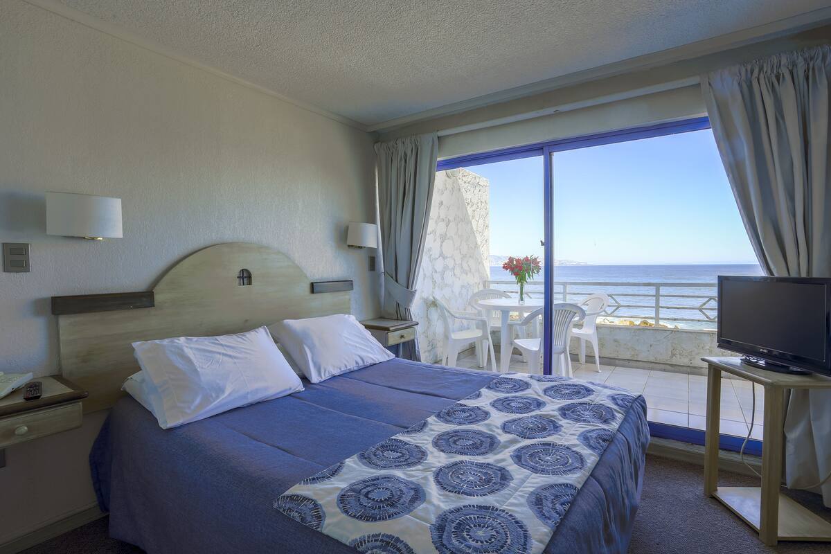 A hotel room with a bed, a TV, a balcony with a table and chairs, and a view of the sea.