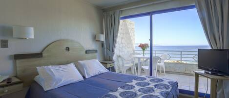 A hotel room with a bed, a TV, a balcony with a table and chairs, and a view of the sea.