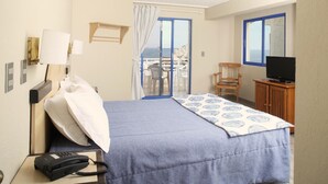A hotel room with a bed, a desk with a telephone, a television, and a view of the sea through a sliding door. - MR Mar Suites (Vina del Mar)