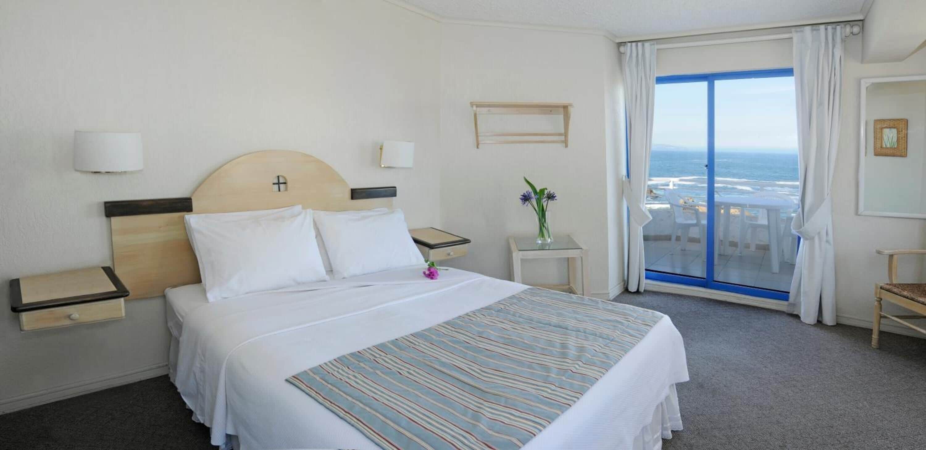 A hotel room with a bed, bedside tables, a small table with a vase, a chair, and a balcony with a view of the ocean.