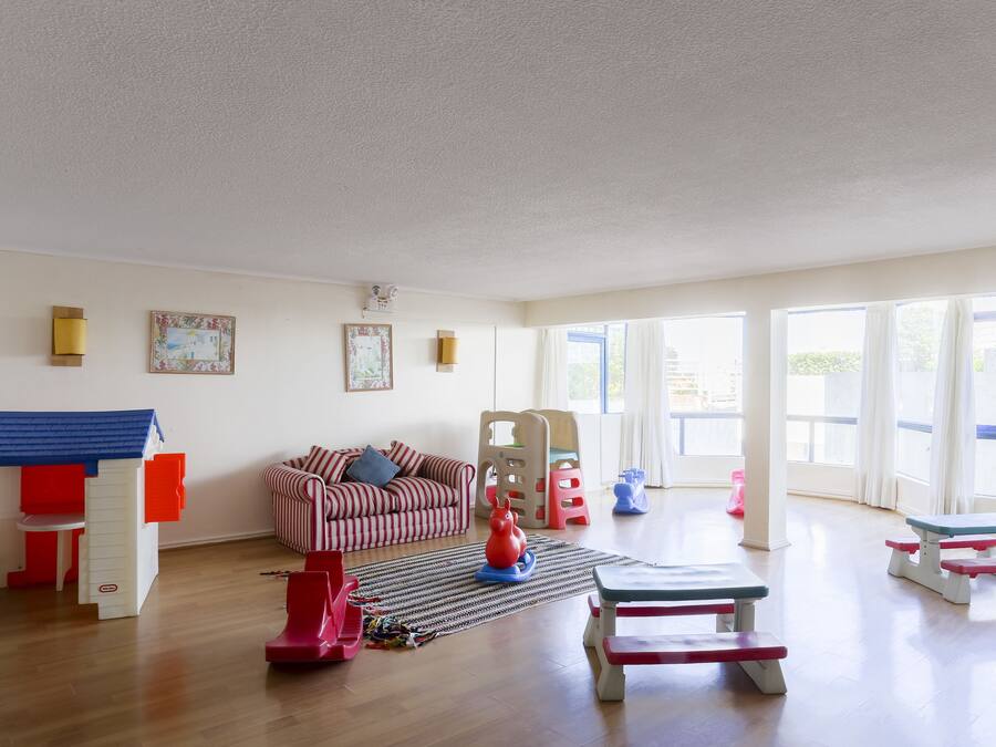 En rummelig stue med trægulv, en rød og hvidstribet sofa, et legeområde med et lille bord og stole, samt store vinduer der lukker naturligt lys ind.
