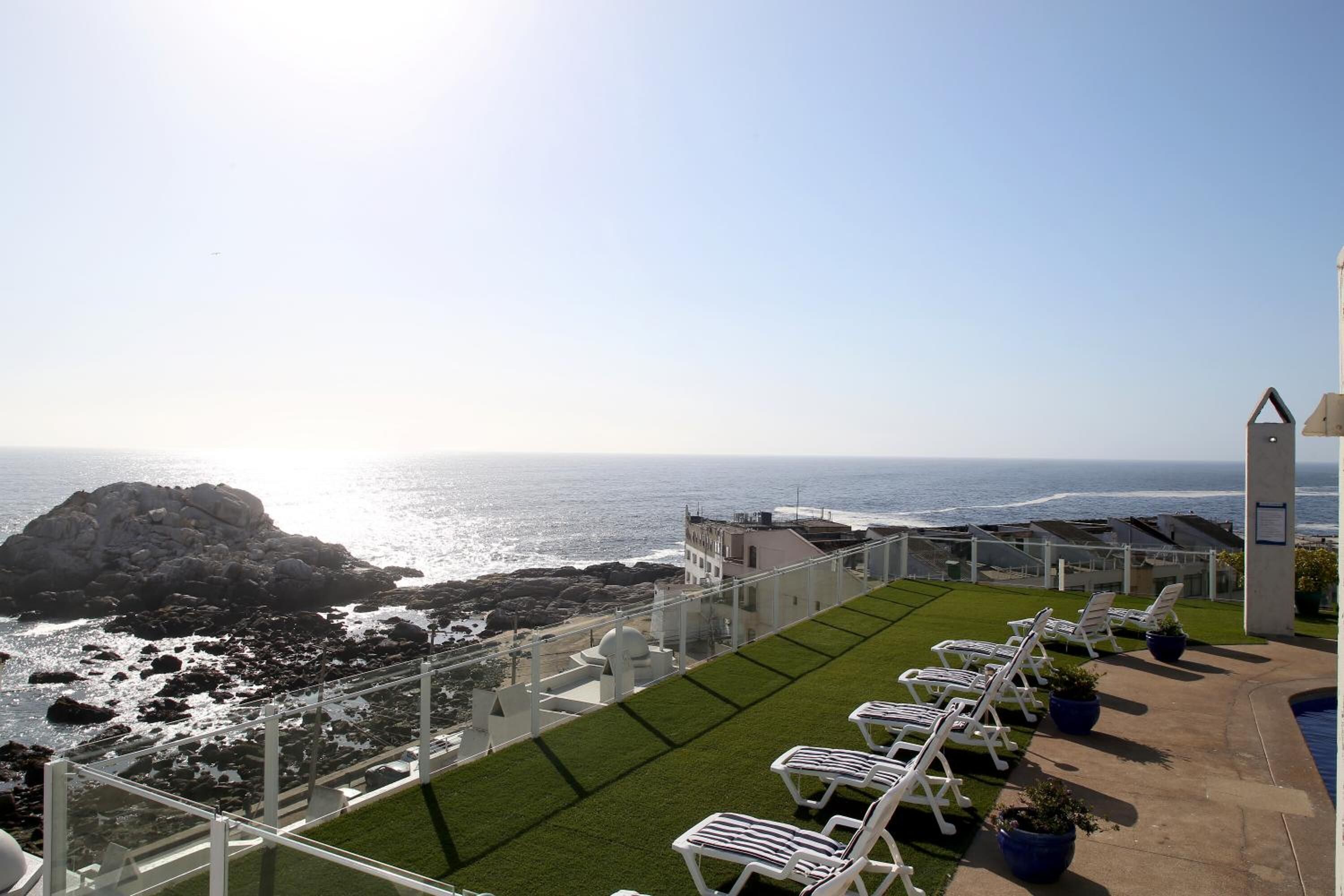 Una terraza con sillones blancos, piscina y vistas al océano y la costa rocosa.