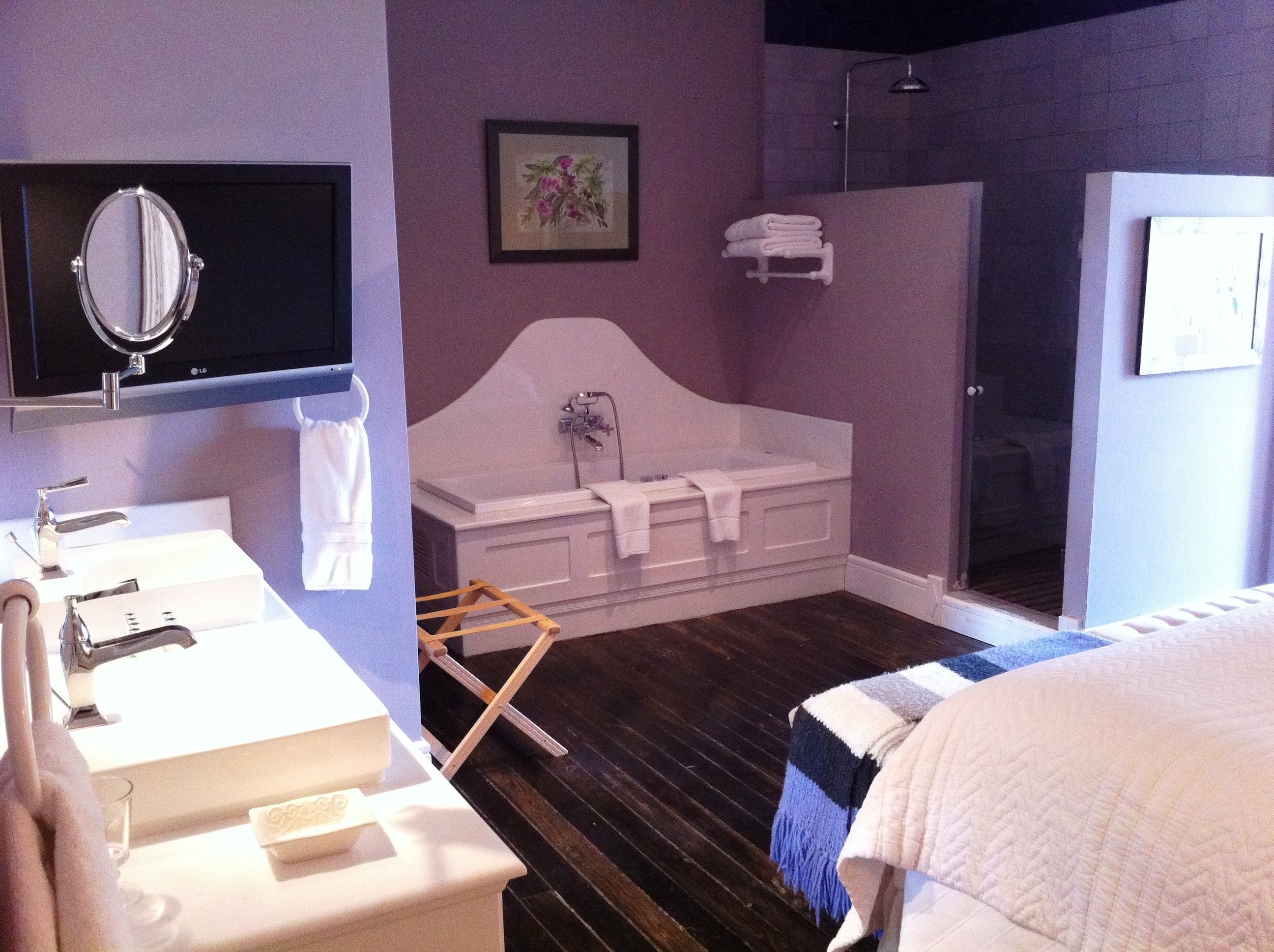 Ein Schlafzimmer mit einem weißen Bett, einer weißen Badewanne, einer Dusche, einem Waschbecken mit Spiegel und Holzboden.