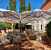 An outdoor patio with wicker chairs and large umbrellas, surrounded by greenery and a building with columns.