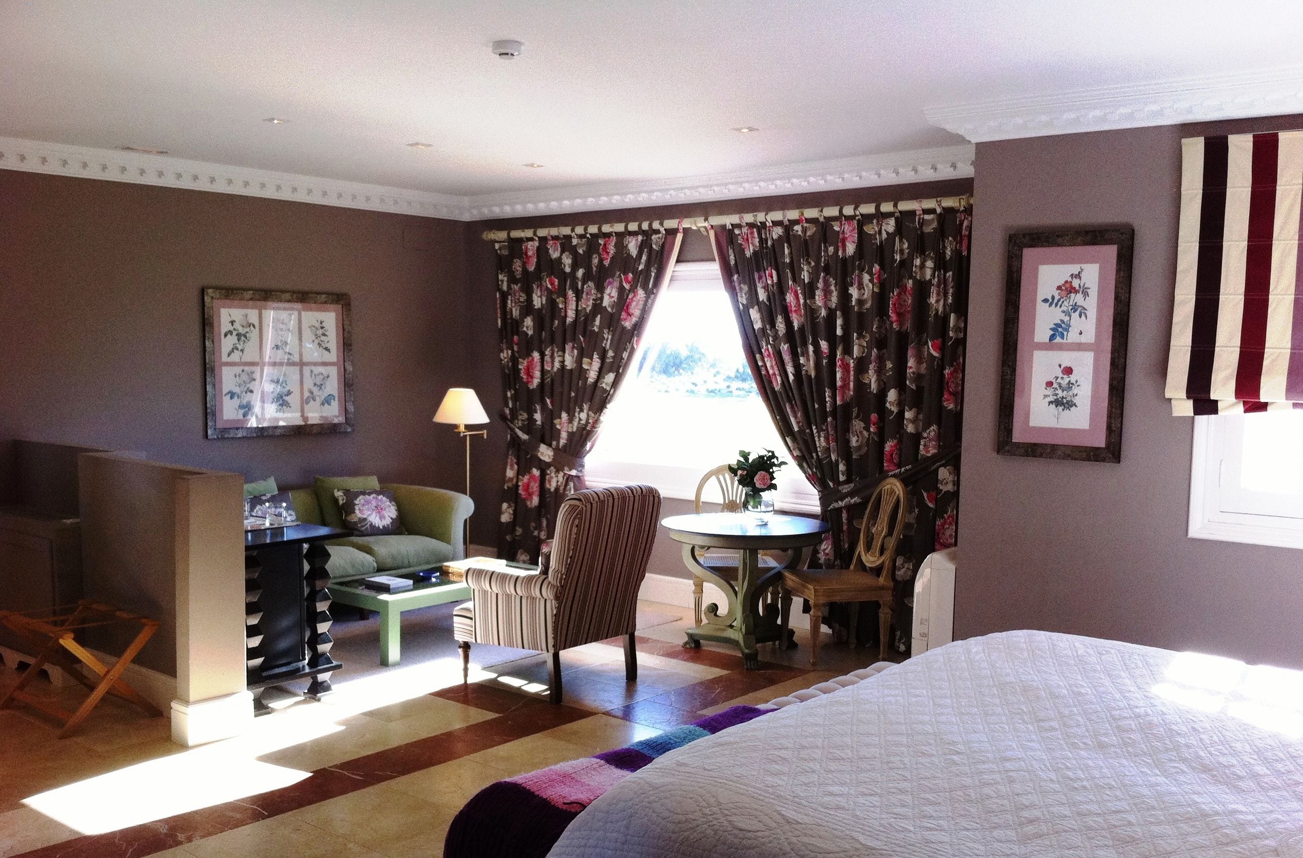 Une chambre d’hôtel comprenant un lit, un bureau, une chaise, une petite table avec un vase de fleurs et une fenêtre avec des rideaux.