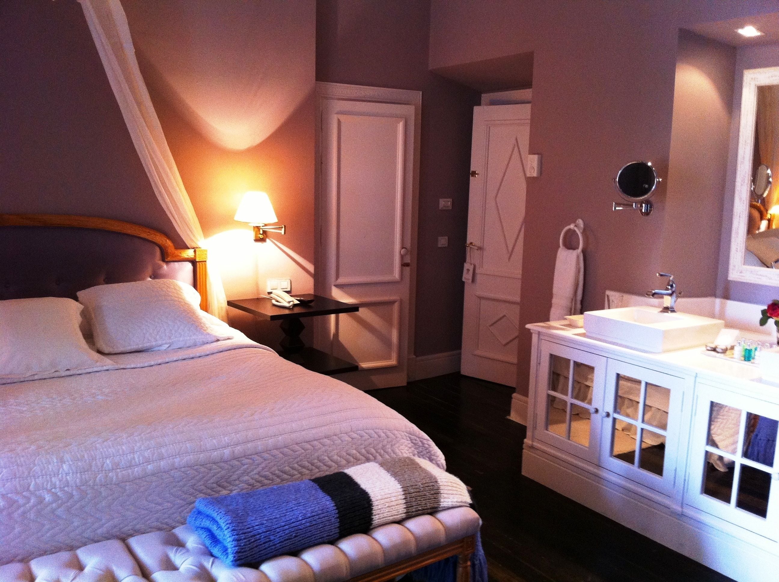 Quarto de hotel com cama, mesa de cabeceira, luminária, guarda-roupa e banheiro com pia e espelho.
