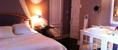 Quarto de hotel com cama, mesa de cabeceira, luminária, guarda-roupa e banheiro com pia e espelho.