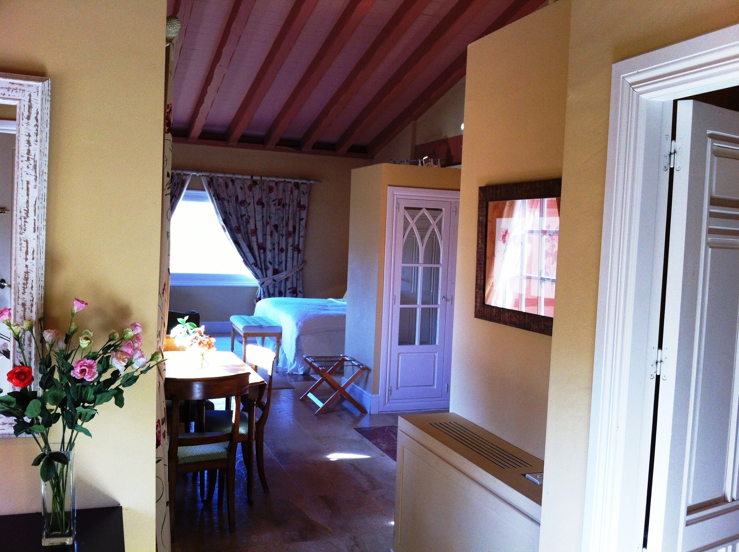 A room with a bed, a table with chairs, a vase of flowers, and a door with a window.