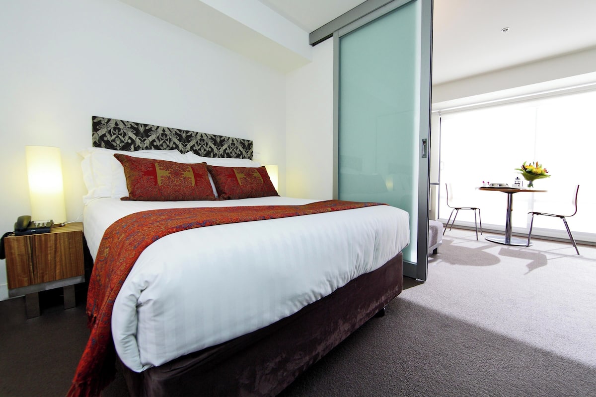 A hotel room with a large bed, a bedside table with a lamp, a small table with a vase of flowers, and a view of the outside through a sliding door.