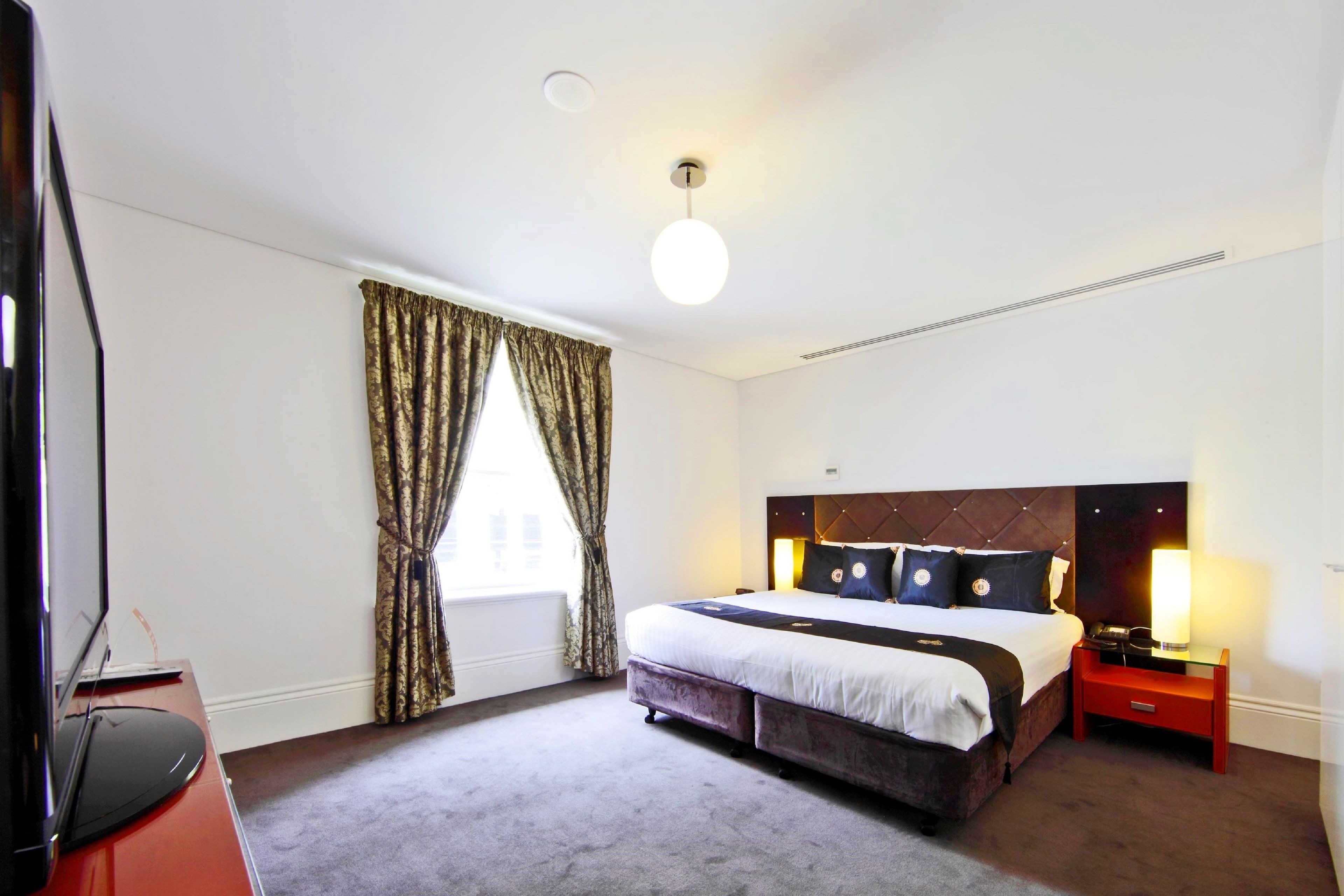 Ein Hotelzimmer mit einem großen Bett, einem Nachttisch mit Lampe, einem Fernseher und einem Fenster mit Vorhängen.