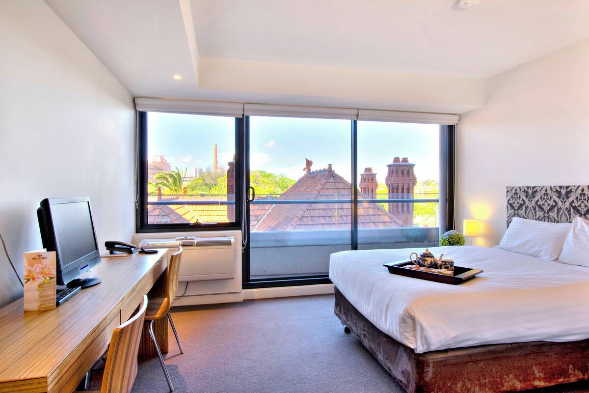 A hotel room with a large window, a bed, a desk with a computer, and a tray with a teapot and cups.