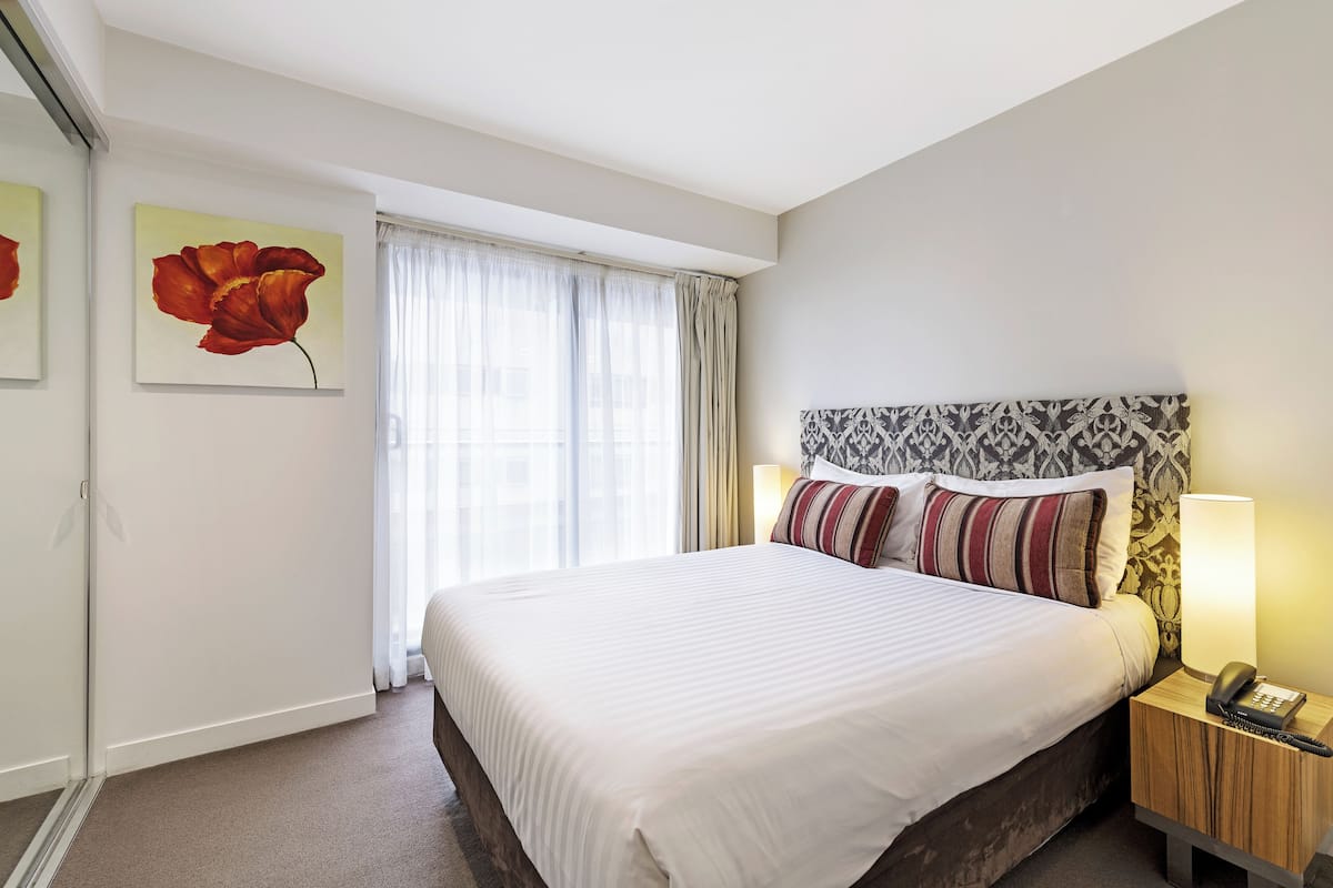 A hotel room with a large bed, a bedside table with a lamp, a telephone, and a window with sheer curtains.