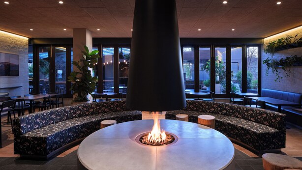 A modern lounge area with a central fire feature, seating arrangements, and large windows providing views of the outdoors. - Hotel Realm (Barton)
