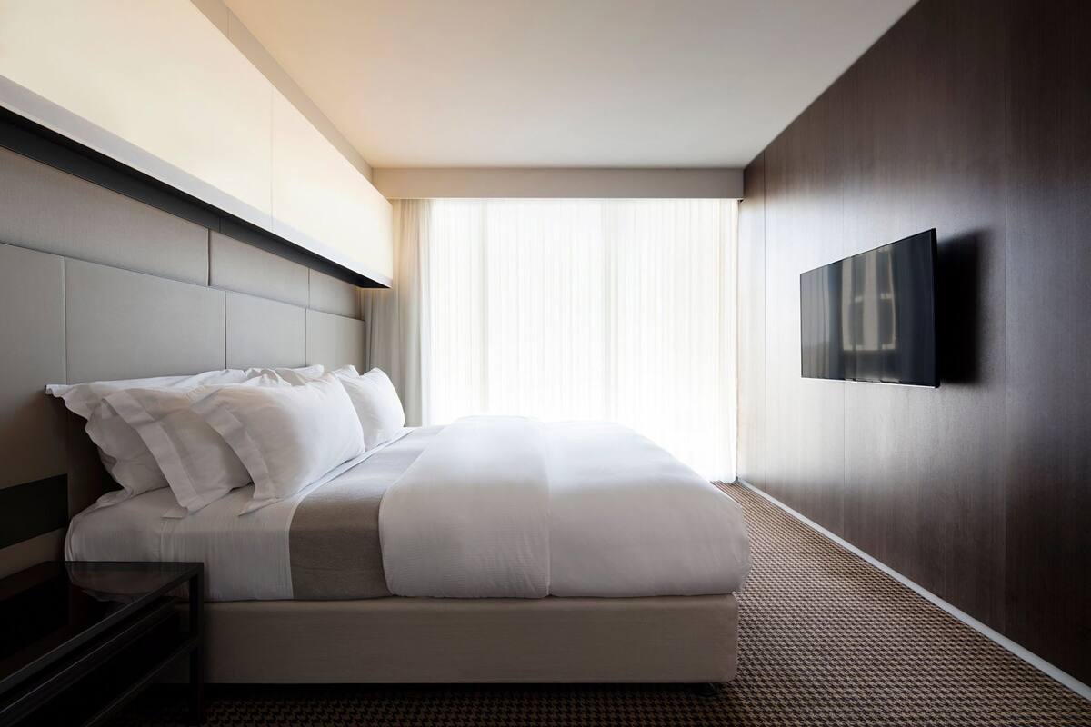 A modern hotel room with a large bed, a flat-screen TV mounted on the wall, and a large window with sheer curtains.